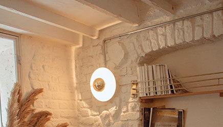 Een gezellige kamer met witgekalkte bakstenen muren en een plafond met houten balken. Een ronde wandlamp is boven een plank gemonteerd, waarop meerdere boeken en kunstwerken van Schilders staan. Gedroogd pampasgras in een vaas is naast een raam geplaatst, waardoor de kamer wordt gevuld met zacht, natuurlijk licht.