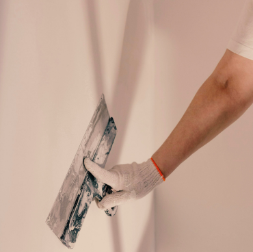 Een persoon met een witte handschoen is te zien terwijl hij pleister of voegmiddel op een muur aanbrengt met een grote metalen troffel, wat de vaardigheid van professionele schilders demonstreert. De achtergrond is een glad, lichtgekleurd oppervlak.