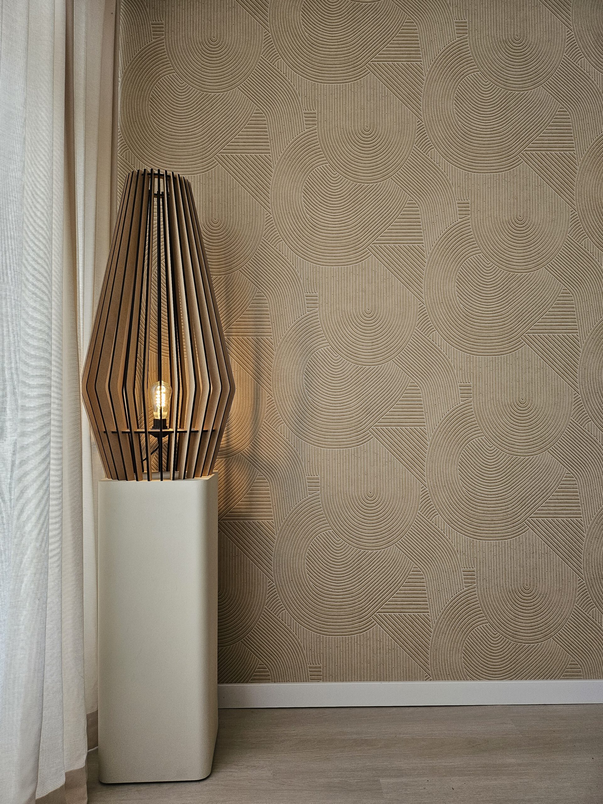 Een moderne houten vloerlamp met een geometrisch kooi-achtig ontwerp staat in een kamer met beige, gepatroneerd behang. De lamp brandt en werpt warm licht op het getextureerde schildersbehang versierd met halve cirkel- en lijnpatronen. Beige gordijnen zijn gedeeltelijk zichtbaar aan de linkerkant.