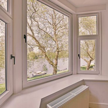 Een lichte, luchtige kamer met drie grote ramen biedt een schilderachtig uitzicht op hoge bomen aan een rivier. De ramen, omlijst in het wit als het werk van bekwame schilders, hebben een witte radiator onder de middelste. Daglicht filtert erdoorheen en benadrukt het rustige buitenlandschap.