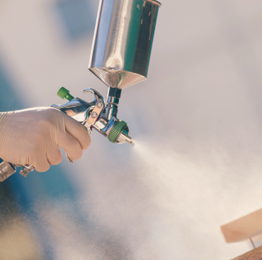 Close-up van een hand met een handschoen die een spuitpistool gebruikt om een fijne nevel verf aan te brengen. De schilderstool heeft een metallic finish met groene accenten en wordt in een hoek gehouden. De achtergrond is zacht vervaagd, waardoor de focus op de tool en de verfnevel wordt gevestigd.