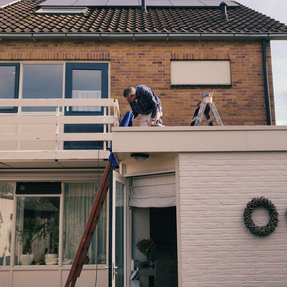Een persoon knielt op het dak van een huis onder een balkon op de tweede verdieping, bezig met schilderen. Ze hebben een doek in hun hand, typisch voor schilders, en er staat een ladder tegen het dak. Het huis is gemaakt van baksteen met een witte gevel en een krans hangt aan de muur. Een persoon knielt op het dak van een huis onder een balkon op de tweede verdieping, bezig met schilderen. Ze hebben een doek in hun hand, typisch voor schilders, en er staat een ladder tegen het dak. Het huis is gemaakt van baksteen met een witte gevel en een krans hangt aan de muur.