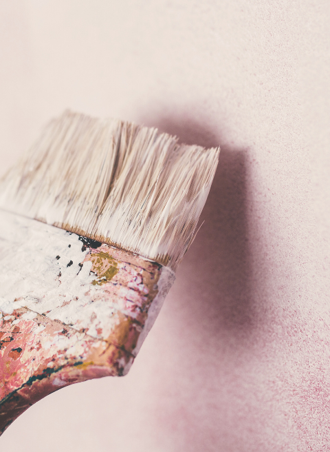 Een close-up van een veelgebruikte schilderskwast met gedroogde verf op het handvat en de haren, die een roze muur borstelt. De zichtbare slijtage van de kwast suggereert dat deze in meerdere schilderprojecten is gebruikt. De achtergrond is zacht vervaagd.