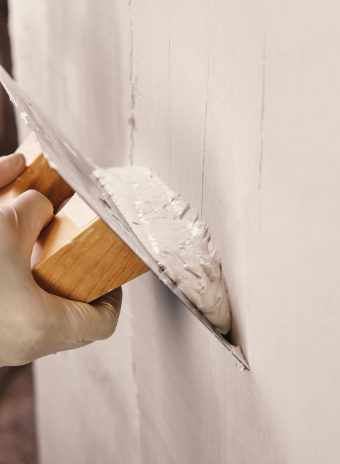 Close-up van een hand met een handschoen die met een troffel pleister op een muur aanbrengt. De muur is gedeeltelijk bedekt met vers pleisterwerk terwijl de persoon, die doet denken aan bekwame schilders, het gladstrijkt. De achtergrond is wazig om de nadruk te leggen op het pleisterproces.