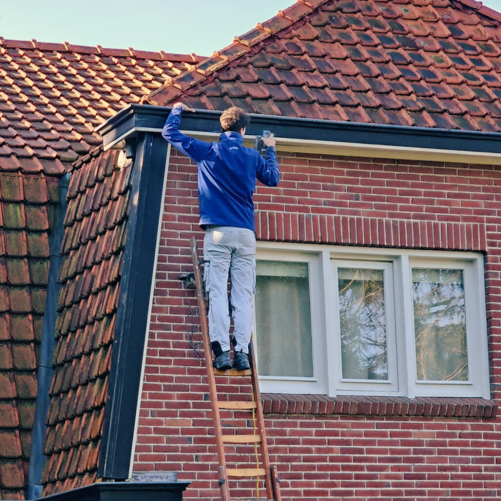 Een persoon in een blauw jasje en een lichtgekleurde broek staat op een ladder en werkt aan de afwerking van het dak van een bakstenen huis. Het huis, waarschijnlijk bijgewoond door schilders, heeft rode dakpannen en een set van twee ramen met witte omlijsting. Daglicht verlicht de scène.