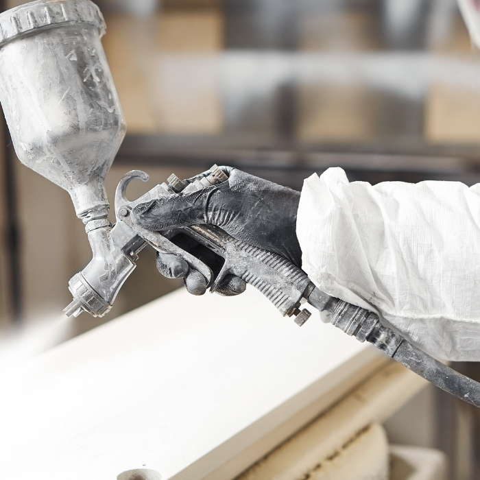 Een persoon die beschermende handschoenen en een wit pak draagt, houdt een spuitpistool vast en brengt een laag verf aan op een plat houten oppervlak in een werkplaats. De achtergrond is wazig, waardoor de aandacht op het schilderproces wordt gevestigd.