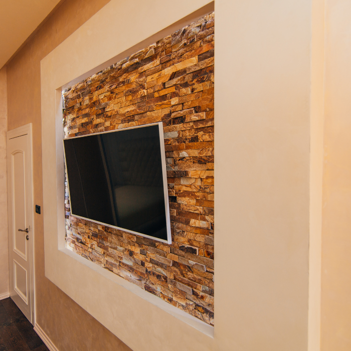 Een flatscreen-tv is aan een muur bevestigd met een decoratieve stenen fineer inleg, omgeven door een beige, getextureerde muur. Een witte deur met een zwarte hendel is gedeeltelijk zichtbaar aan de linkerkant. De opstelling is onderdeel van een modern interieur kamerontwerp.