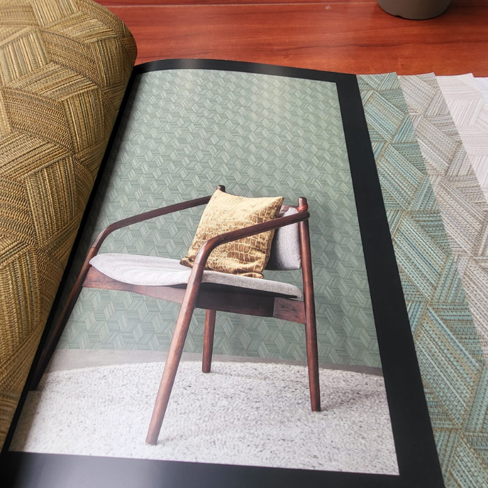 Een cataloguspagina toont een moderne houten stoel met een beige kussen en een mosterdgeel sierkussen tegen een lichtgroene muur met geometrisch patroon. De catalogus ligt op een houten oppervlak en toont gedeeltelijk aangrenzende stofstalen in verschillende patronen en kleuren.