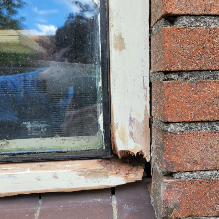 Close-up van een beschadigd raamkozijn naast een bakstenen muur. Het houten frame vertoont tekenen van rot en afbladderende verf bij de onderste hoek. Een weerspiegeling van een persoon is zichtbaar in het glas en een deel van een airconditioningunit is door het raam te zien.