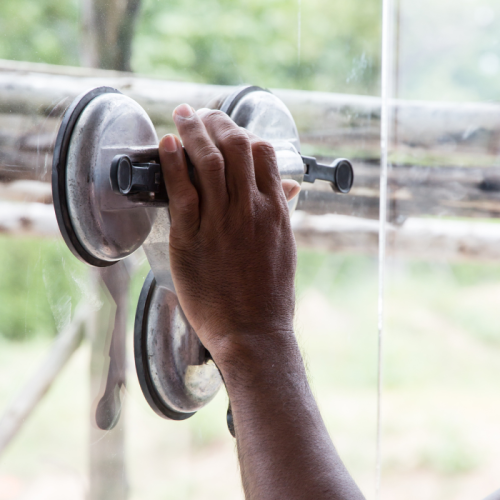 Een close-up van de hand van een persoon die een dubbele zuignap vasthoudt die aan een glazen oppervlak is bevestigd. De achtergrond toont een buitenscène met wazig groen en houten structuren.
