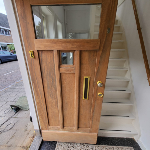 Een houten voordeur met een klein rechthoekig raampje bovenaan en een brievenbus van messing eronder. De deur heeft verticale paneelontwerpen en messing beslag, waaronder een deurkruk en een slot. De deur opent naar een gang met een witte trap en een houten leuning. Buiten is er een straat en een buurhuis.