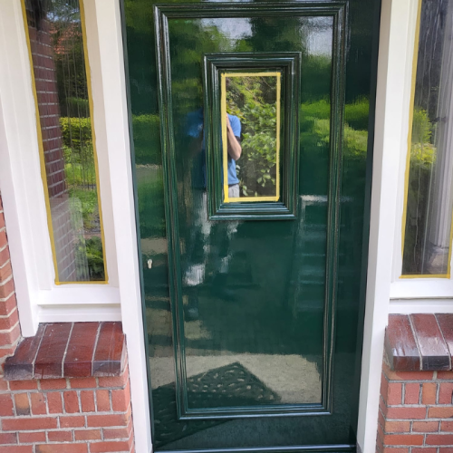 Een glimmende groene voordeur met een rechthoekig raam in het midden, omlijst door gouden versieringen. De deur is omgeven door een wit frame en rode bakstenen muren. De weerspiegeling van een persoon die de foto neemt is zichtbaar in het raamglas. Groen is zichtbaar op de achtergrond.