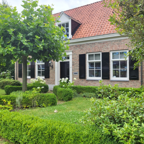 Een charmant bakstenen huis met een rood pannendak, zwarte luiken en grote ramen. Een goed onderhouden tuin met weelderige groene heggen, struiken en een boom in de voortuin. De lucht is gedeeltelijk bewolkt.