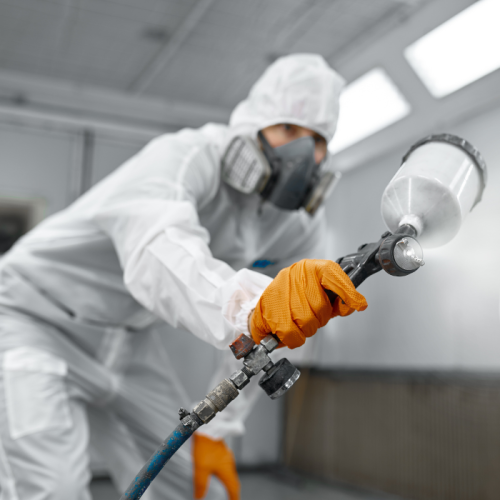 Een persoon in beschermende kleding en handschoenen spuit verf met een spuitpistool in een industriële omgeving. De nadruk ligt op de veiligheid door een ademhalingsmasker te dragen en de aandacht te richten op de bediening van de apparatuur.