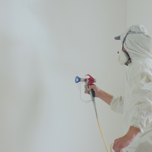 Een persoon in beschermende uitrusting, waaronder een kap, masker en handschoenen, gebruikt een verfspuit om een witte coating op een muur aan te brengen. De kamer lijkt in het proces van schilderen te zijn, met minimale zichtbare meubels of apparatuur.