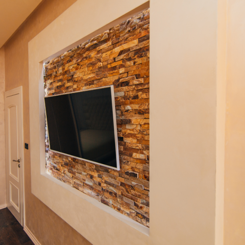 Een flatscreen-tv is aan een muur bevestigd met een decoratieve stenen fineer inleg, omgeven door een beige, getextureerde muur. Een witte deur met een zwarte hendel is gedeeltelijk zichtbaar aan de linkerkant. De opstelling is onderdeel van een modern interieur kamerontwerp.