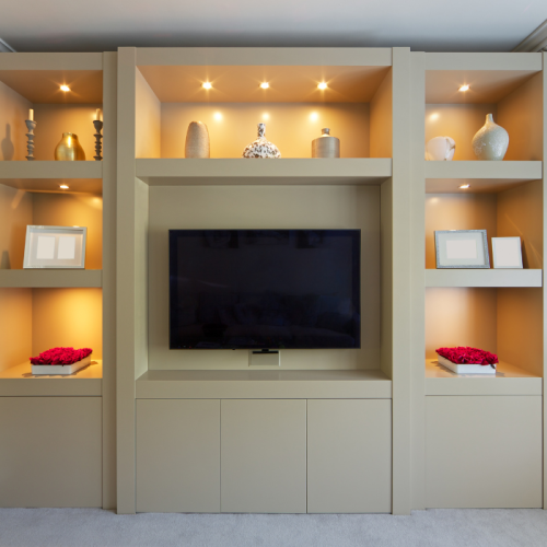 Een moderne woonkamer-entertainmentunit met een grote flatscreen-tv in het midden. De omringende planken zijn versierd met verschillende items, waaronder vazen, ingelijste foto's en kommen gevuld met rode bloemen. Geïntegreerde verlichting benadrukt het decor.