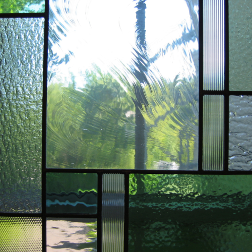 Een close-up van een glas-in-loodraam met verschillende getextureerde en gepatroneerde doorschijnende panelen in tinten groen en helder. Zonlicht filtert door het glas, wat reflecties en een wazig zicht op bomen en een pad buiten werpt.