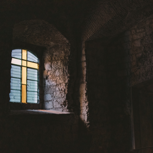 Een klein, gebogen raam met een geel kruisontwerp van glas-in-lood werpt een zacht licht in een verder donkere kamer met stenen muren. De textuur van de oude stenen muren en de zachte verlichting creëren een serene en historische sfeer.